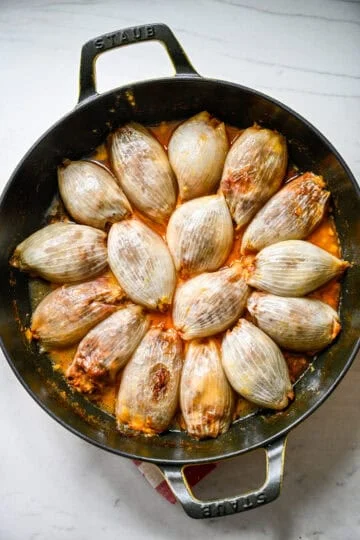 Overhead view of baked stuffed onions in a Staub cast iron skillet, golden brown on top and sitting in broth