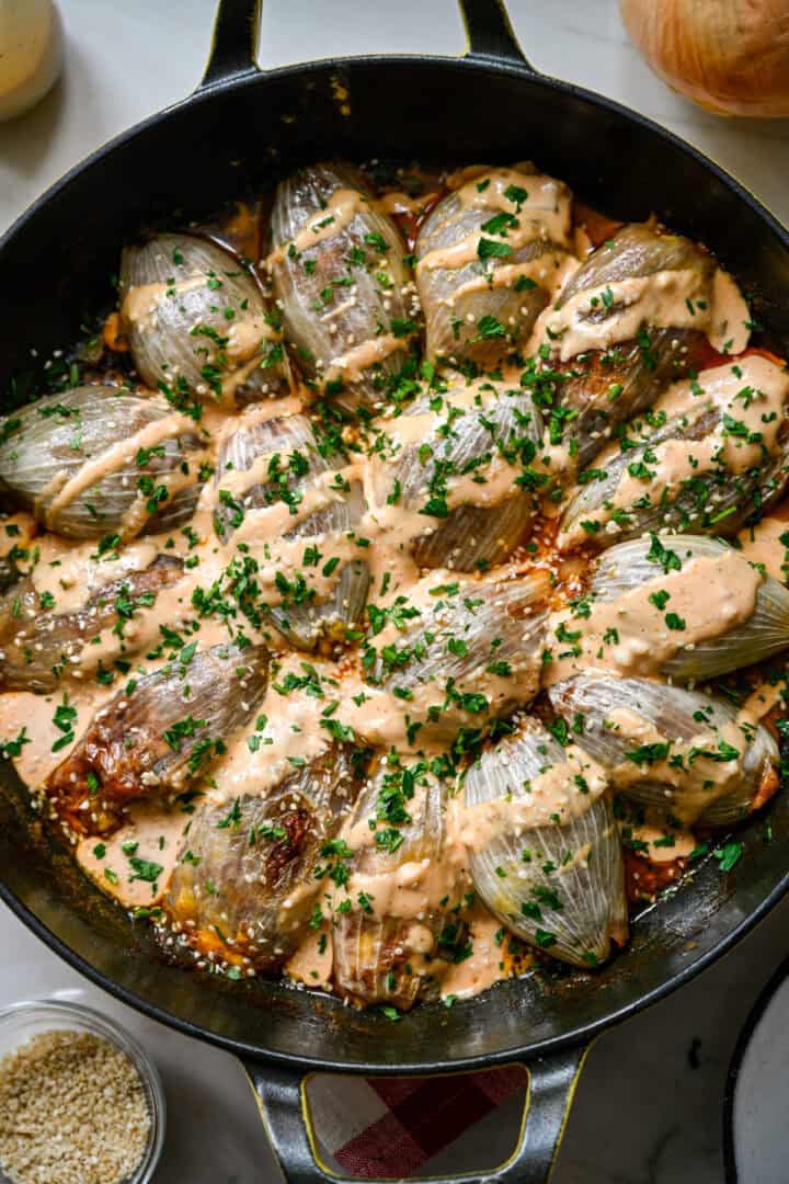 45 degree view of a skillet of bacon cheeseburger stuffed onions topped with Thousand Island dressing, parsley and sesame seeds on a marble counter