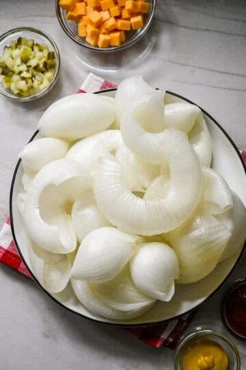 Overhead view of boiled onion layers peeled apart and piled on a white plate, with bowls of chopped pickles and cubed cheddar