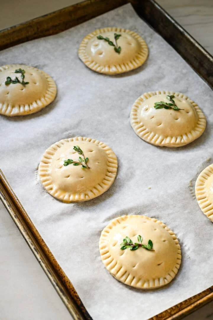 45 degree view of six unbaked hand pies on a parchment-lined sheet pan, brushed with egg wash and topped with thyme sprigs and steam holes.
