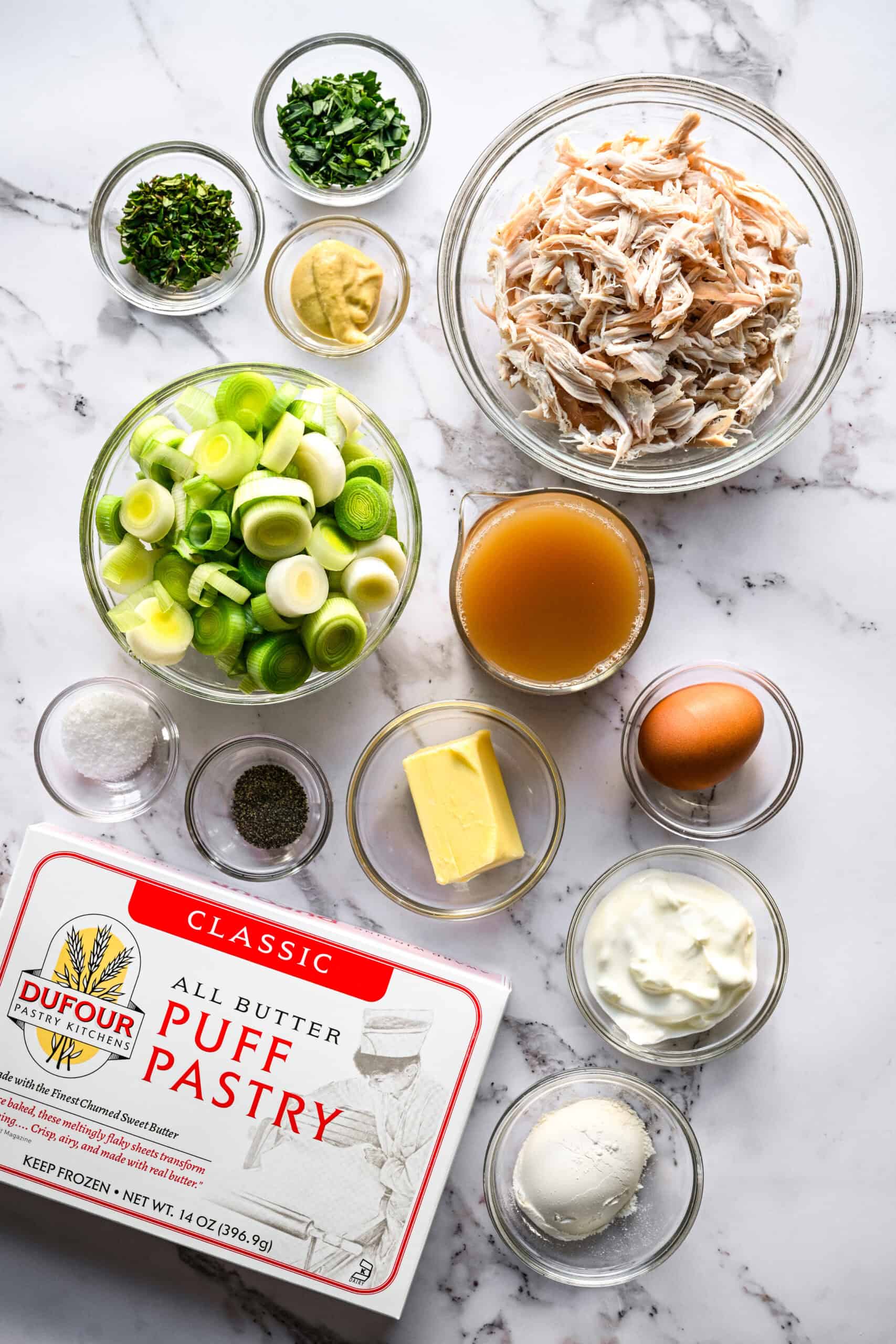 Overhead view of ingredients for chicken and leek hand pies on marble: shredded chicken, sliced leeks, chicken stock, a box of Dufour all butter puff pastry, butter, sour cream, flour, an egg, Dijon, tarragon, thyme, salt and pepper in glass bowls.