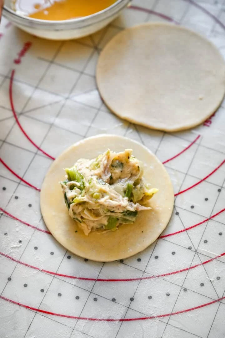 45 degree view of a mound of chicken and leek filling on a round of puff pastry on a pastry mat, with a second pastry round and a bowl of egg wash behind.
