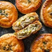 Overhead view of a chicken and leek hand pie cut in half to show the creamy chicken and leek filling and flaky layers, surrounded by whole hand pies on a dark tray.