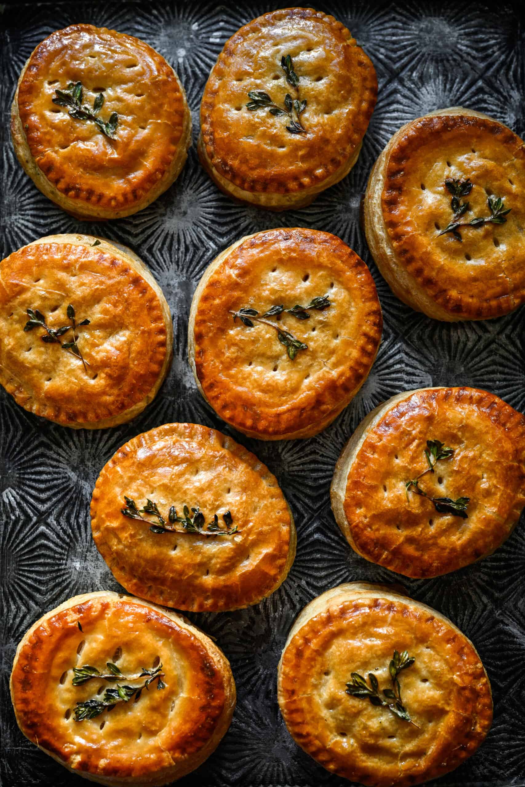 Overhead view of ten baked chicken and leek hand pies with thyme sprigs on a dark patterned metal tray.