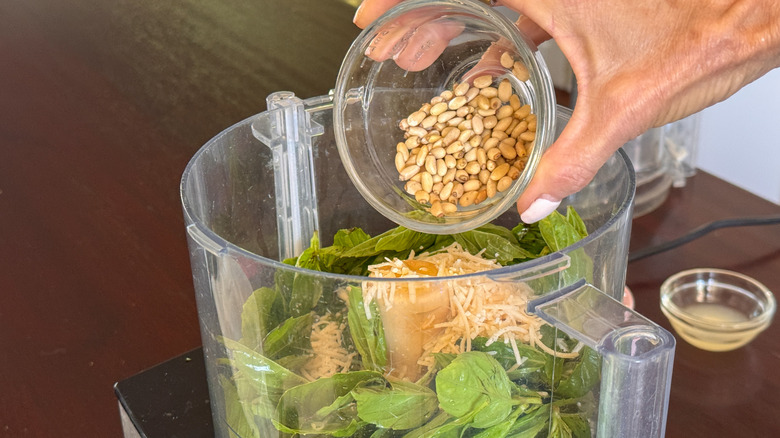 hand adding pine nuts to food processor full of basil