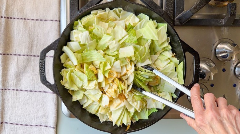 Using tongs to stir chopped green cabbage in skillet on stovetop