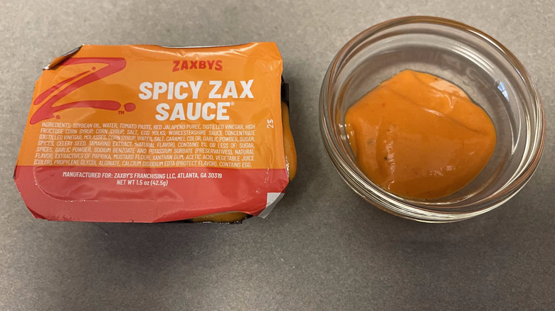 A packet of Spicy Zax Sauce sits on a gray counter next to a dish of orange sauce.