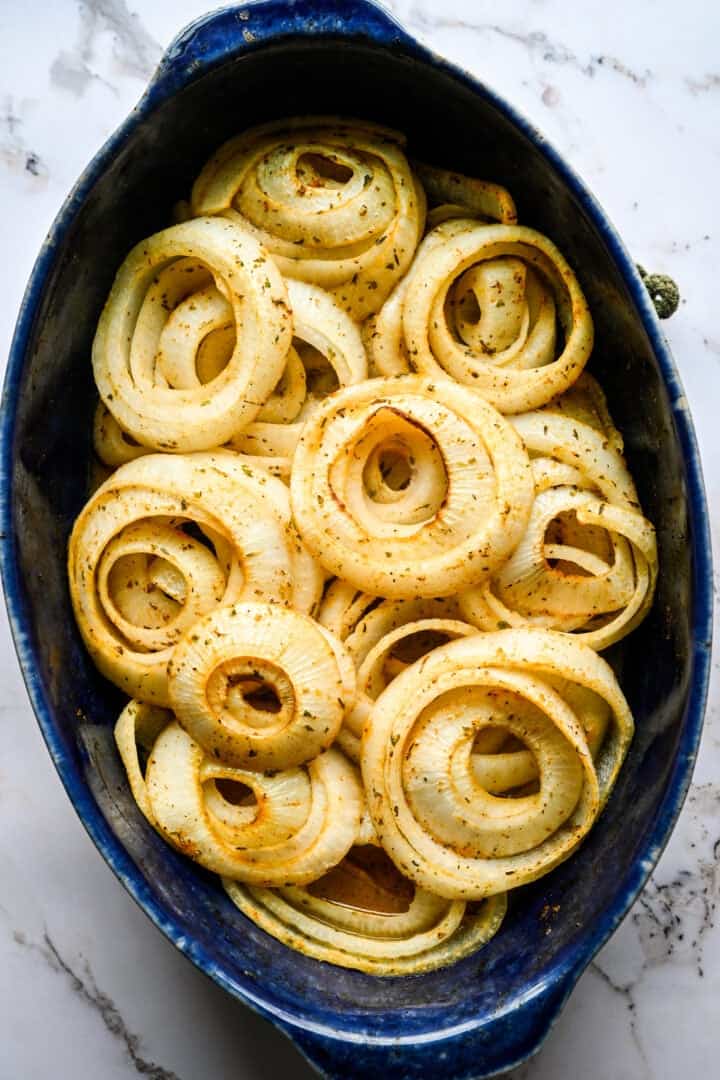 Overhead view of seasoned raw onion rings in a blue oval baking dish, with smaller rings nested inside larger ones.