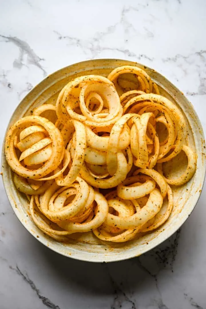 Overhead view of raw sweet onion rings in a wide ceramic bowl after shaking, evenly coated in paprika and dried herbs.