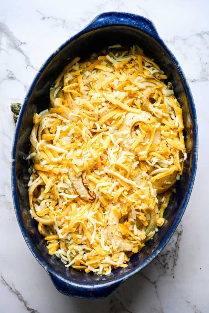 Overhead view of partially roasted onion rings in a blue oval baking dish, topped with shredded mozzarella and cheddar before the final bake.