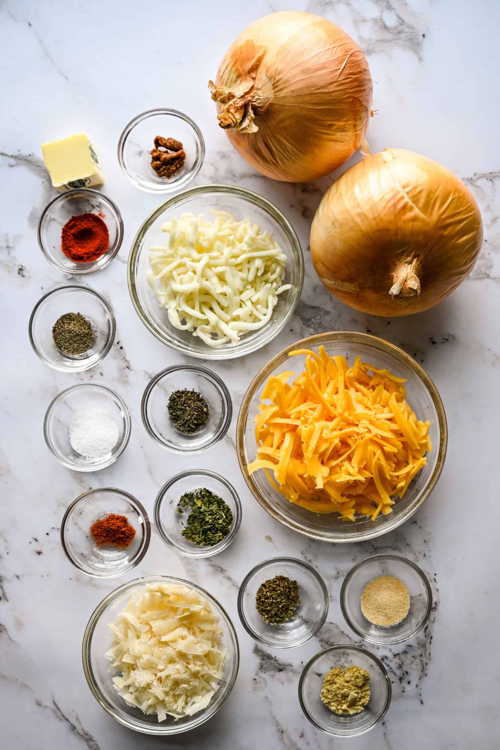 Overhead view of ingredients for Tennessee onions on a marble surface: two whole sweet onions, butter, bowls of shredded mozzarella and cheddar, grated Parmesan, dried herbs and spices.