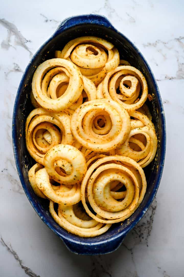 Overhead view of seasoned raw onion rings arranged in a blue oval baking dish, ready to go into the oven.